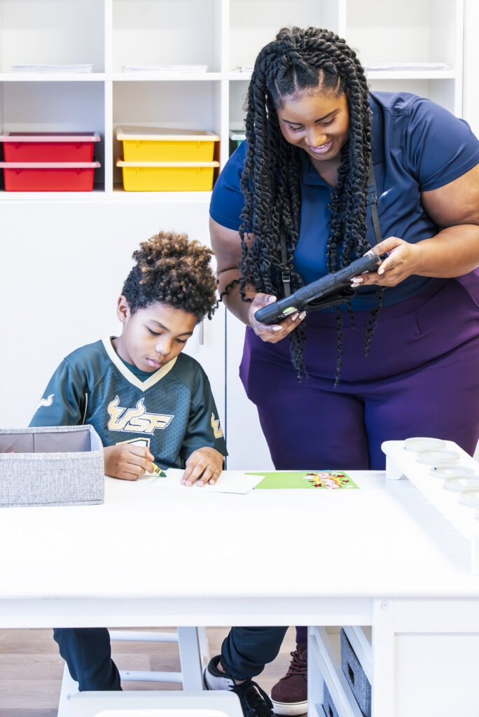 Child sitting at desk coloring, with woman standing next to him looking at a tablet