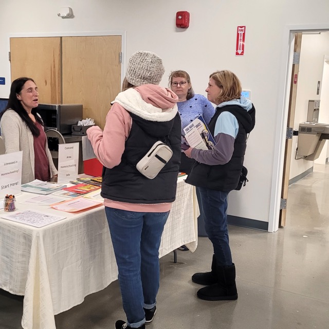 Visitors discuss homeschooling at a table with books, handouts, and a Concord-area homeschooling sign.