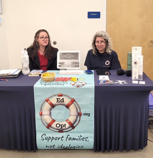 Two EdOpt representatives sit behind an information table with brochures, giveaways, and a lifebuoy banner.