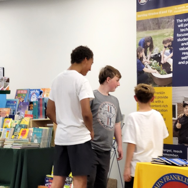 Three young visitors talk beside the Benjamin Franklin Academy table and a children’s book display.