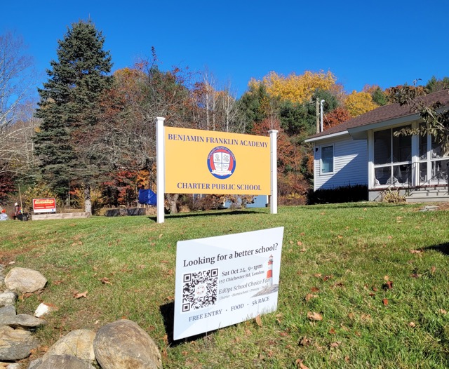 Benjamin Franklin Academy’s roadside sign stands behind an education fair lawn sign amid autumn foliage.