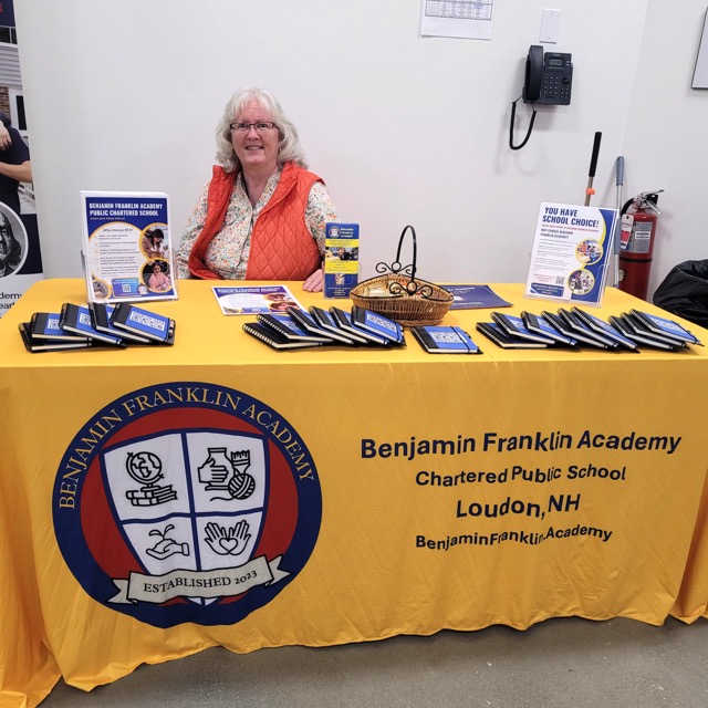 A Benjamin Franklin Academy representative sits behind a yellow table with brochures and school-choice information.