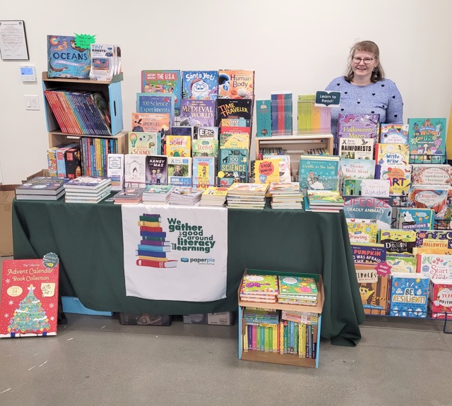 A PaperPie representative stands behind a display of children’s books, activity books, and reading resources.