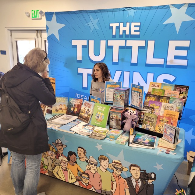 A visitor browses books and talks with a representative at a colorful Tuttle Twins display.