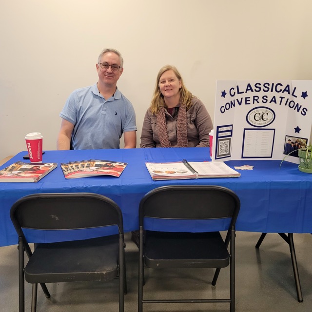 Two Classical Conversations representatives sit behind a table with brochures and an information display.