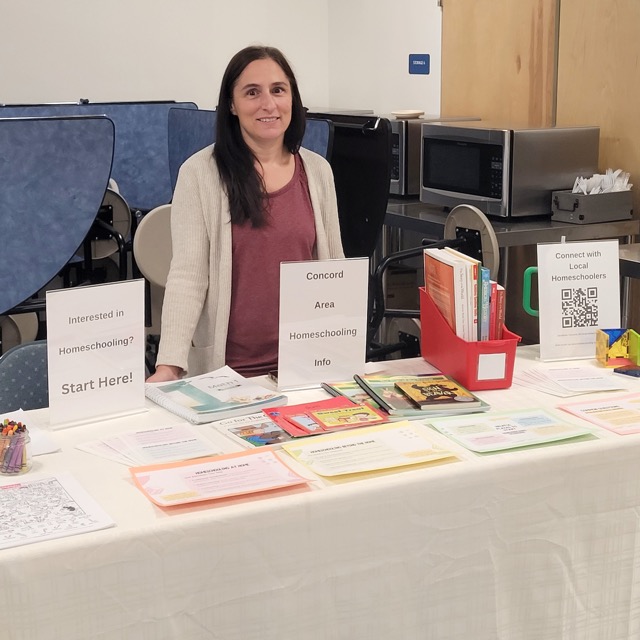 A representative stands behind a Concord-area homeschooling table with books, handouts, and a local connections QR code.