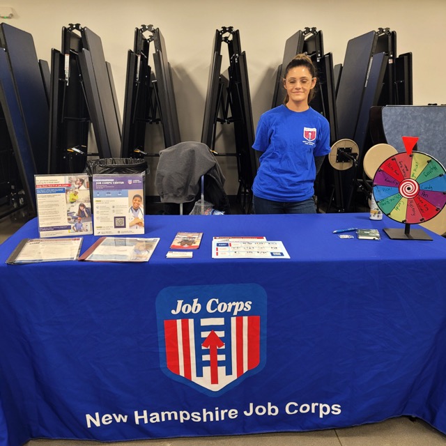 A New Hampshire Job Corps representative stands behind a blue table with brochures and a colorful prize wheel.