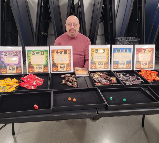 A man sits behind a dice probability game with candy, dice trays, and signs showing the odds of different totals.