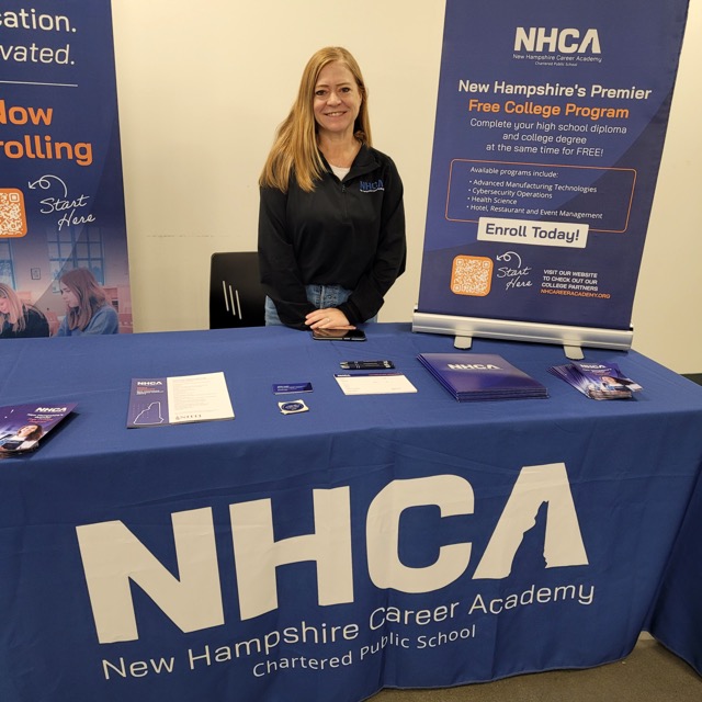 A New Hampshire Career Academy representative stands behind a blue information table beside a free-college-program banner.